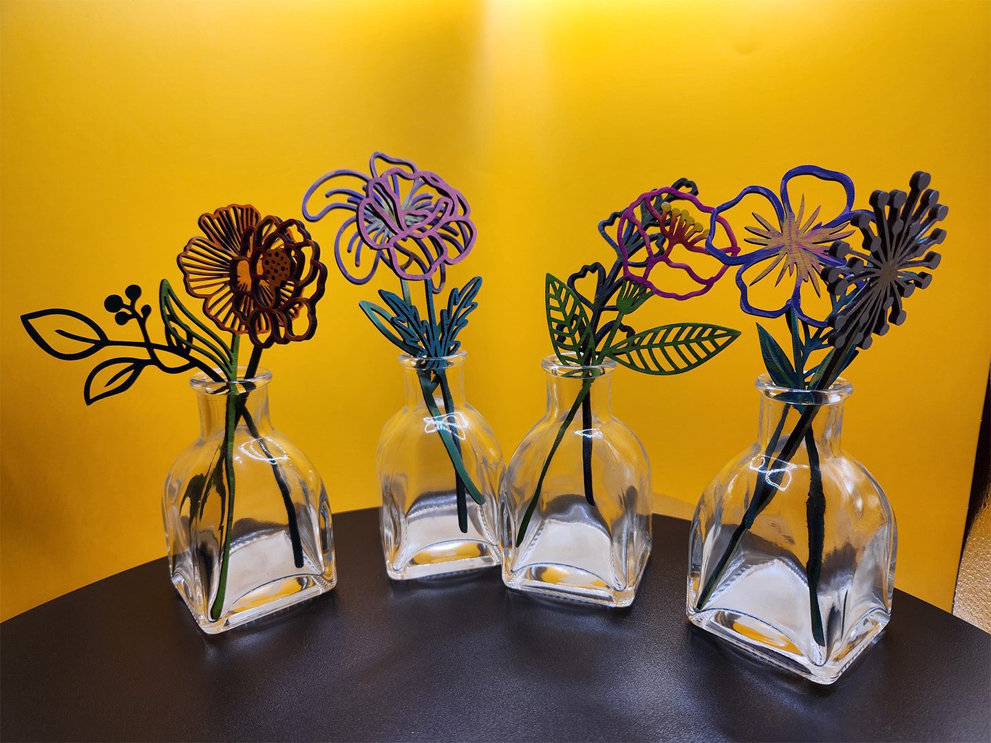 Four glass bottles each holding a colorful wire flower against a yellow background.